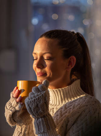 Beautiful girl with knitted gloves holding cup of hot coffee beside window