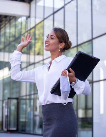 Attractive business woman holding laptop and facial protective mask and waving hand in front od office buildingの写真素材