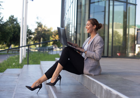 Pretty young business woman sitting on stair in front of glass building, holding laptop in lap and having conference callの写真素材