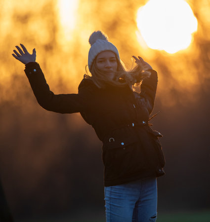Pretty teenage girl in jacket and with cap jumping cheerful in park at sunset in winter timeの写真素材