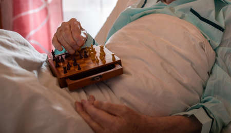 Close up of senior man's hand holding chess board and pieces in lap in medical bed in hospitalの写真素材