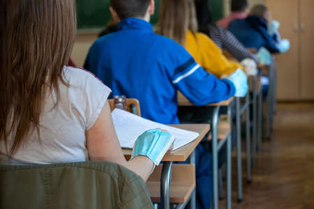 Rear view of pupils sitting at desks in classroom with protective masks on hands. Pandemic protection conceptの写真素材