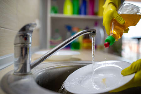Close up of female hands with gloves pouring liquid detergent on plate for dishwashing under tap waterの写真素材
