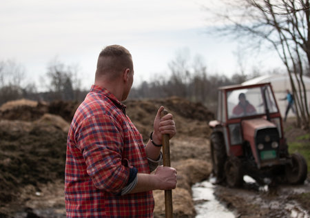 Rear view of farmer with hayfork standing in pile of natural manure with tractor in background