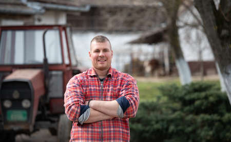 Happy farmer with crossed arms standing in front of tractor on farm