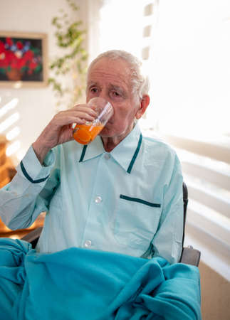 Senior man sitting in wheelchair covered with blanket and drinking juice beside window at homeの写真素材