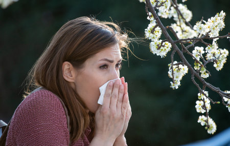 Pretty young girl blowing nose in front of blooming tree. Spring pollen allergy attack conceptの写真素材