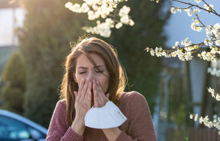 Young pretty woman sneezing in front of blooming tree. holding protective mask in hand. Spring pollen allergy attackの写真素材