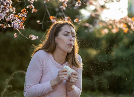 Pretty young woman sneezing in front of blooming tree. Spring allergy attack conceptの写真素材