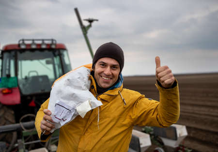Portrait of handsome farmer carrying sack with seeds for sowing field on shoulder to tractorの写真素材