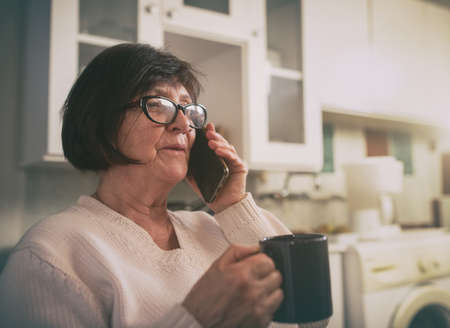 Senior woman holding cup of coffee and talking on mobile phone in kitchen at homeの写真素材