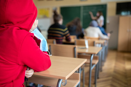 Rear view of pupils sitting at desks in classroom with protective masks on hands. Pandemic...