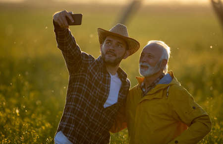 Two farmers standing in field taking selfie using phone smiling happy. Men marking in agribusiness...