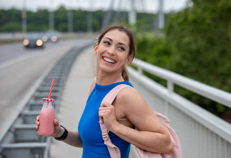 Young athlete smiling laughing walking on bridge. Fit woman having strawberry milkshake smoothie...