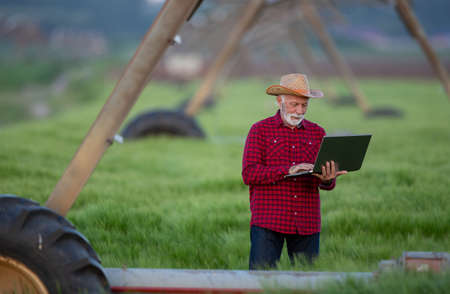 Senior farmer holding laptop and standing under irrigation systemの写真素材