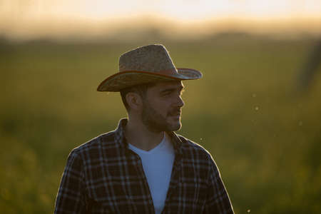 Portrait of young man farmer with straw hat standing in field at sunset in springtimeの写真素材