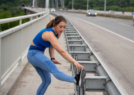 Young fit woman stretching leg on bridge railing after exercise. Flexible athlete exercising...