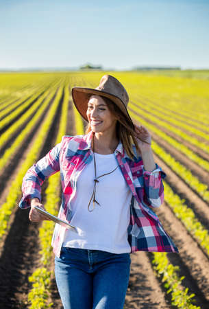 Young female agronomist standing in soy field smiling. Farmer using tablet in agriculutre holding hat looking to side.の写真素材