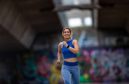 Athlete running smiling in front of wall with graffiti. Girl wearing sportswear exercising.