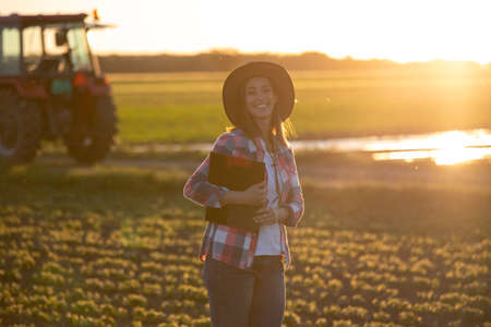 Young woman farmer looking at camera in field holding clipboard. Agronomist standing on plantation in front of tractor machinery.の写真素材