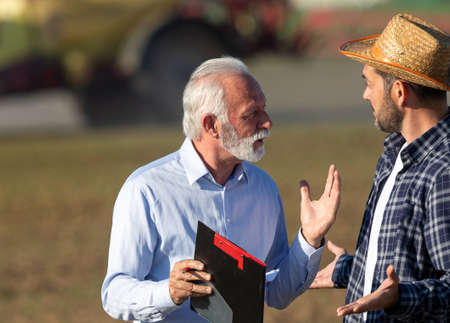 Two men talking arguing explaining in field with machinery in background. Farmer and insurance sales...