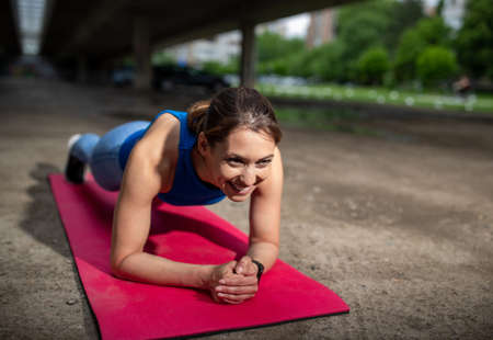 Female athlete exercising outdoors in parking lot. Fit young woman doing plank on pink mat smiling.
