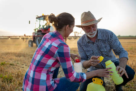 Farmer man and woman pouring chemicals for plants care into bottle in front of tractor with sprayer in fieldの写真素材