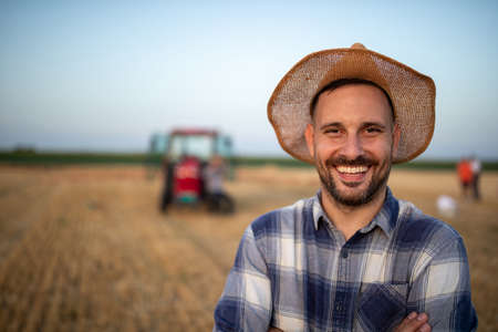Portrait of satisfied handsome farmer with straw hat on head in field during harvest with tractor in backgroundの写真素材