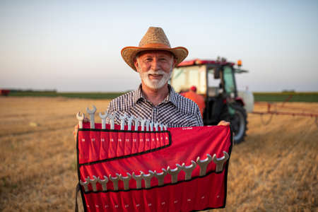 Portrait of senior farmer holding set of wrenches in front of tractor in fieldの写真素材