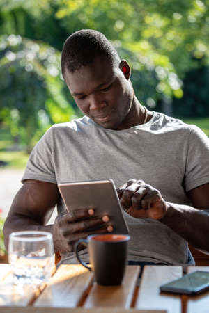 Attractive Afro American man relaxing in garden with drink, using tablet technology while sitting at table.の写真素材