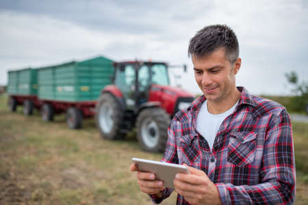 Male agronomist looking at camera smiling while using tablet. Handsome farmer standing in harvested field in front of tractor with trailer.の写真素材