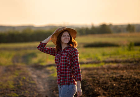 Attractive young woman wearing plaid shirt standing in field looking back. Ginger girl farmer holding sunhat on head smiling.の写真素材