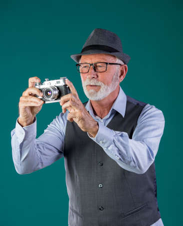 Senior man with white beard, in vintage clothes and hat holding old analog camera on blue background in studioの写真素材