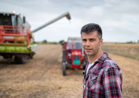 Attractive young agronomist standing in harvested field looking at camera. Combine harvester filling tractor trailer with grainsの写真素材