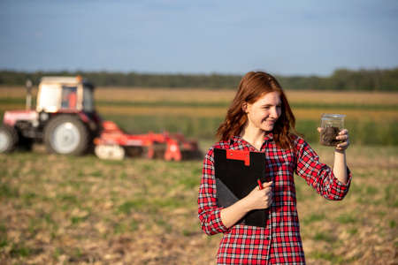 Young woman agronomist standing in field holding soil sample. Attractive young farmer monitoring land quality with tractor working behind.の写真素材
