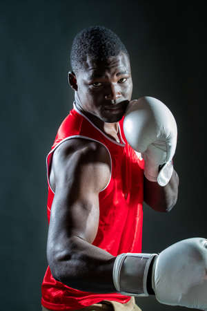 Young man with dark skin boxing in jersey with gloves against blue gray background in studioの写真素材