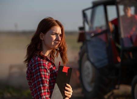 Pretty female agronomist standing in field taking notes using clipboard. Male farmer sitting in tractor with door open in background.の写真素材