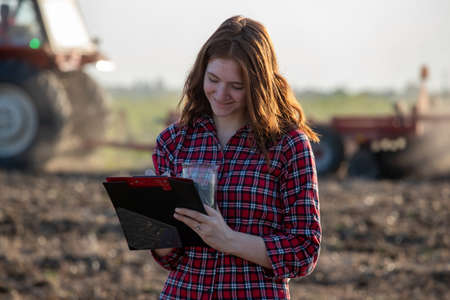 Attractive female agronomist holding soil sample in beaker writing in clipboard. Young woman smiling standing outdoors with tractor working land behind.の写真素材