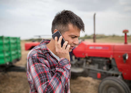 Attractive young farmer talking on phone while standing outdoors in harvested field in front of tractor with trailer in background.の写真素材