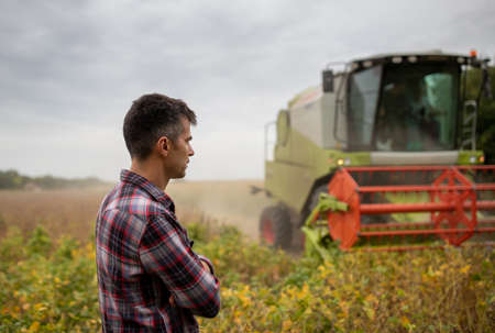 Handsome agronomist standing with crossed arms in soy field checking monitoring crops in early autumn with combine harvester working in backgroundの写真素材