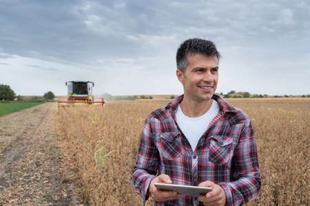 Male agronomist smiling and using tablet modern technology in agriculture. Man standing in soy field with harvester working in background.の写真素材