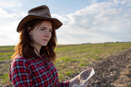 Portrait of young farmer woman with hat and plaid shirt standing in soy field in summer time, holding tablet and monitoring crop growthの写真素材