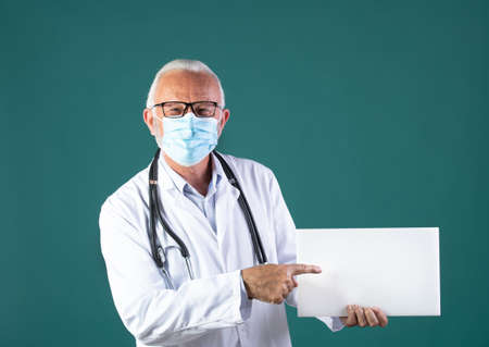 Portrait of senior doctor in white coat with stethoscope around neck and protective mask holding and showing blank white board, against blue backgroundの写真素材