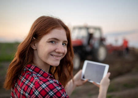 Attractive ginger girl standing in field with tractor in background smiling. Portrait of female farmer holding tablet looking at camera.の写真素材