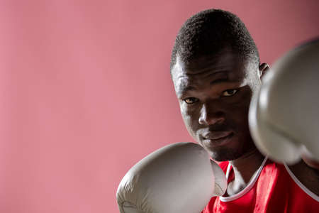 Young man with dark skin boxing in jersey with gloves against pink background in studioの写真素材