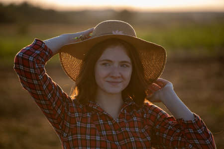 Attractive redhead farmer smiling at sunset. Portrait of girl standing in field holding sunhat.の写真素材