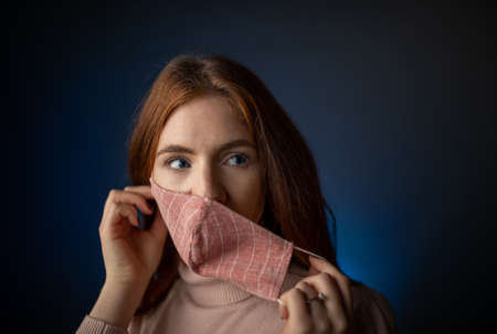 Portrait of pretty young woman with red hair and blue eyes putting protective face mask on, in studio with dark backgroundの写真素材