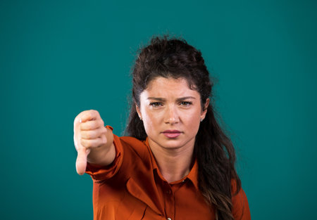 Pretty young woman showing thumb down as negative sign, hand gesture, in studio, against blue backgroundの写真素材