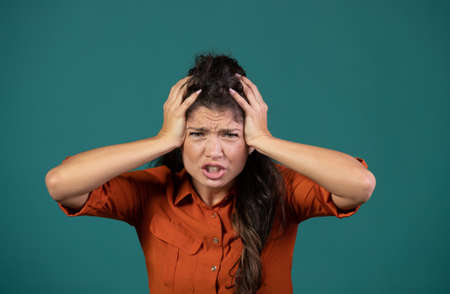 Frustrated young woman holding hands on head, madness concept, in studio on blue background