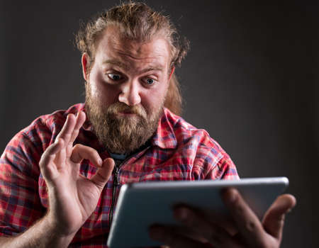 Man with beard and long hair wearing plaid shirt, holding tablet and showing ok sign, satisfied...